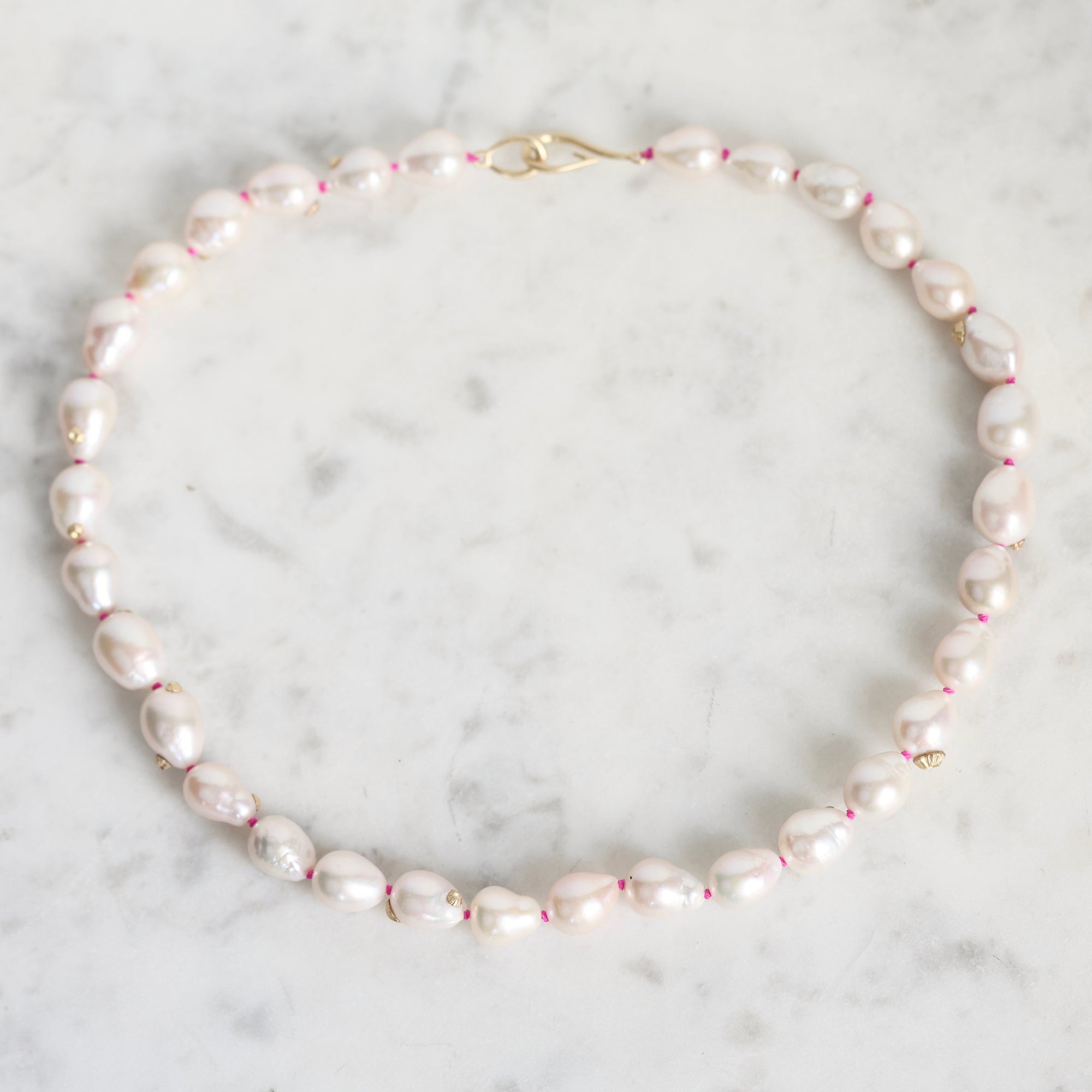 A baroque pearl necklace with a gold clasp, laid flat on a white marble surface. The pearls are light pink and white, separated by tiny pink spacers, and accented with small gold beads.