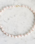 A baroque pearl necklace with a gold clasp, laid flat on a white marble surface. The pearls are light pink and white, separated by tiny pink spacers, and accented with small gold beads.