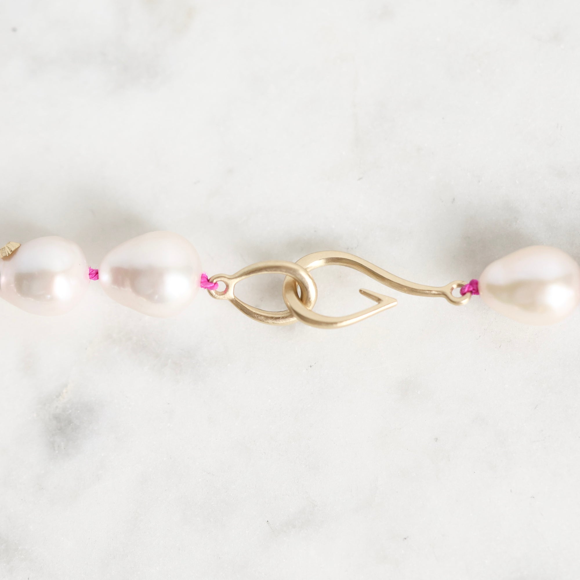 A close-up of a pearl necklace with a gold clasp on a light marble surface. The necklace features large, irregular white pearls separated by small pink accents, with a polished gold S-shaped clasp.