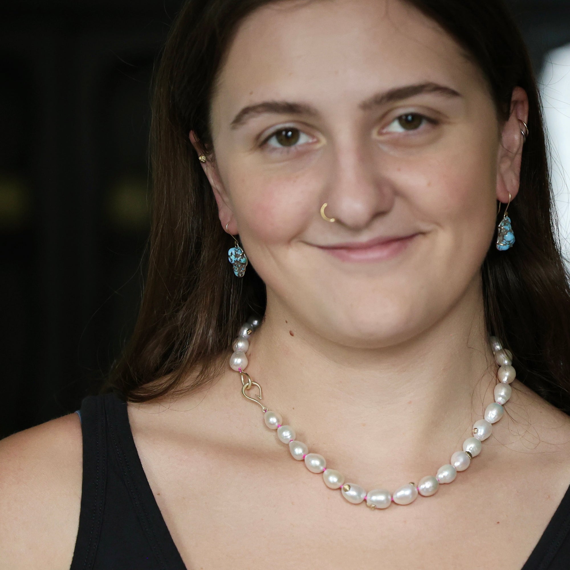 A close-up portrait of a woman wearing a baroque pearl necklace with a gold clasp and matching blue teardrop earrings.