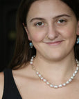 A close-up portrait of a woman wearing a baroque pearl necklace with a gold clasp and matching blue teardrop earrings.