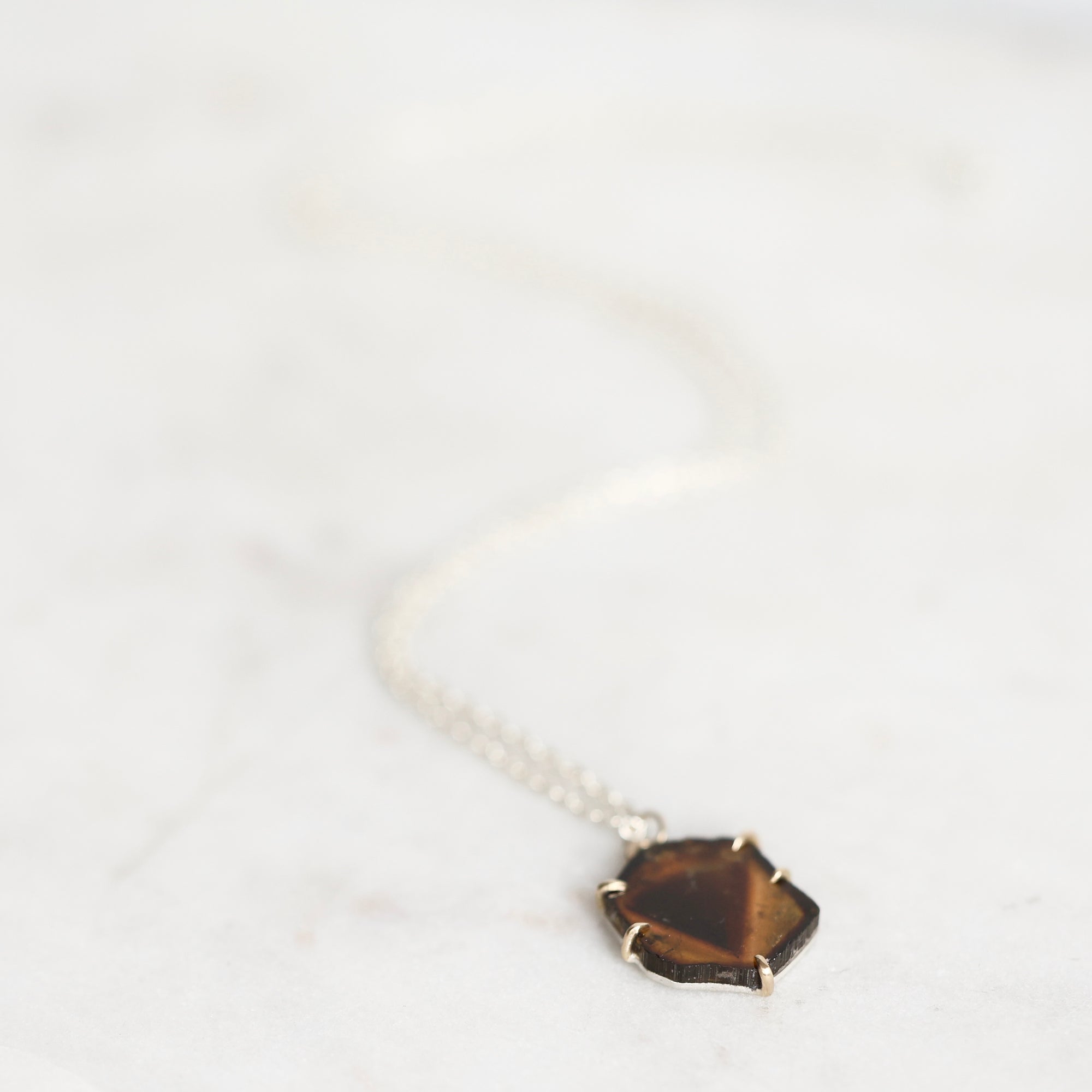 A delicate silver chain necklace with a small, irregular faceted brown gemstone pendant, resting on a light marble surface.