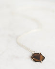 A delicate silver chain necklace with a small, irregular faceted brown gemstone pendant, resting on a light marble surface.