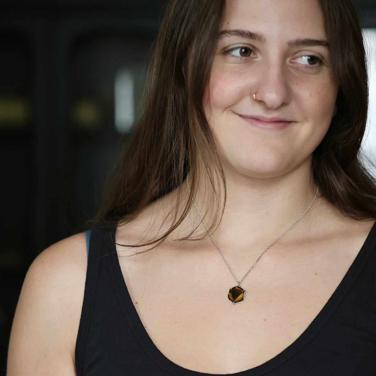 A close-up lifestyle shot of a woman with long brown hair wearing a silver chain necklace with a small, faceted amber-brown pendant, set against a dark, blurred background.