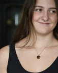 A close-up lifestyle shot of a woman with long brown hair wearing a silver chain necklace with a small, faceted amber-brown pendant, set against a dark, blurred background.