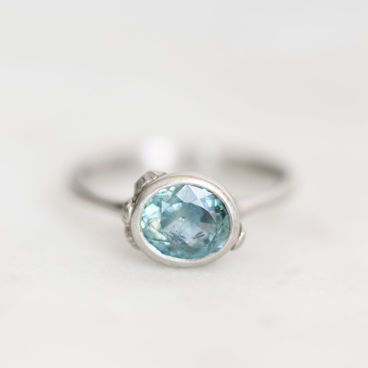 A silver ring featuring an oval-cut light blue gemstone in a bezel setting, photographed on a clean, light off-white background.