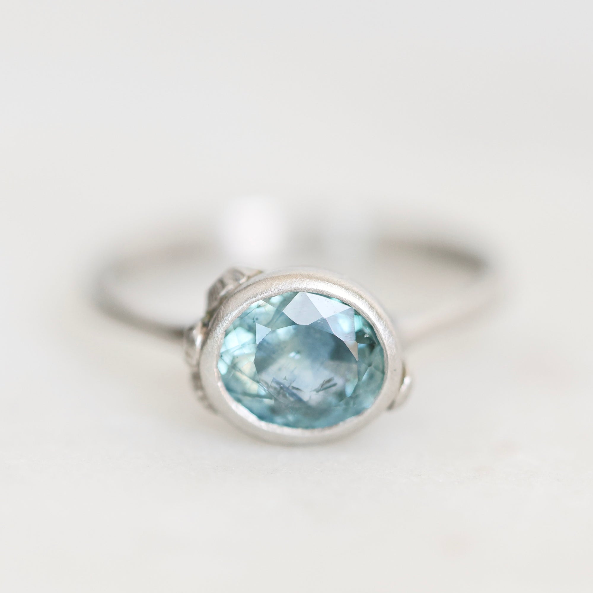 A silver ring featuring an oval-cut light blue gemstone in a bezel setting, photographed on a clean, light off-white background.