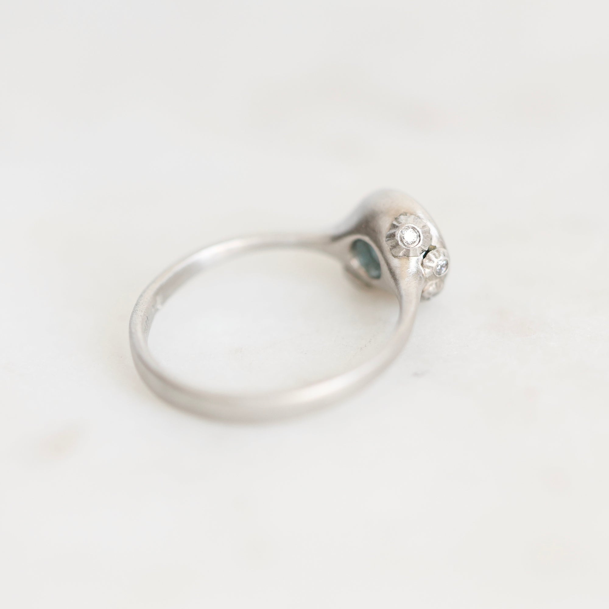 A silver ring with a decorative head featuring small clear stones and a tiny blue-green accent, photographed on a light neutral background.