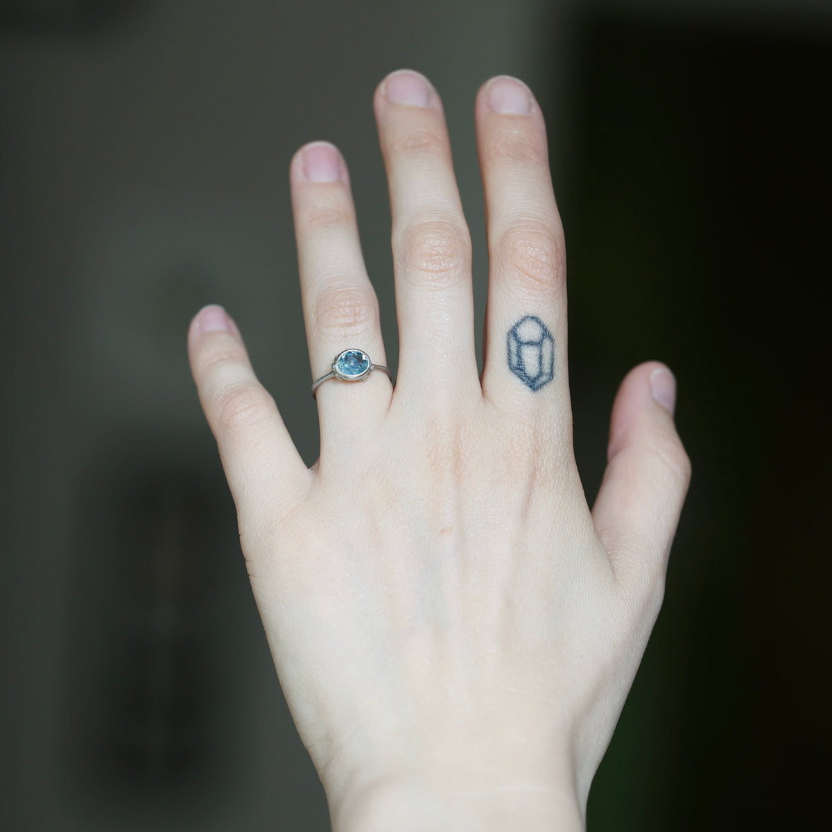 A close-up of a person's hand showing two rings: a silver band with a round light blue gemstone on the ring finger, and a small silver geometric ring on the middle finger. The background is a dark, blurred gray.