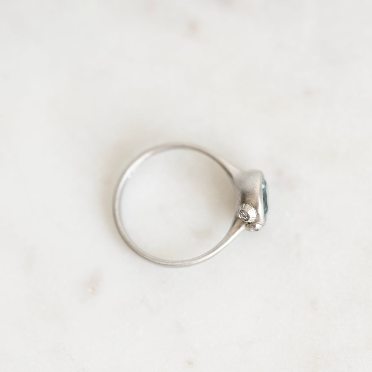 A silver ring with a small oval blue gemstone and a tiny clear accent stone, photographed on a light marble surface.