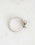 A silver ring with a small oval blue gemstone and a tiny clear accent stone, photographed on a light marble surface.