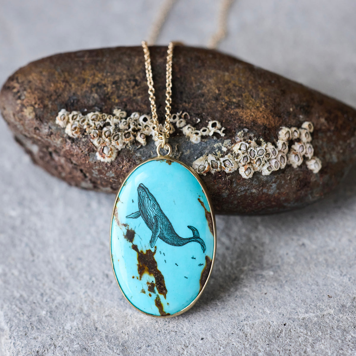 An oval turquoise pendant necklace with a black whale illustration and brown matrix details, hanging from a gold chain in front of a textured rock.