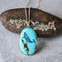 An oval turquoise pendant necklace with a black whale illustration and brown matrix details, hanging from a gold chain in front of a textured rock.