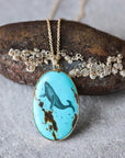 An oval turquoise pendant necklace with a black whale illustration and brown matrix details, hanging from a gold chain in front of a textured rock.