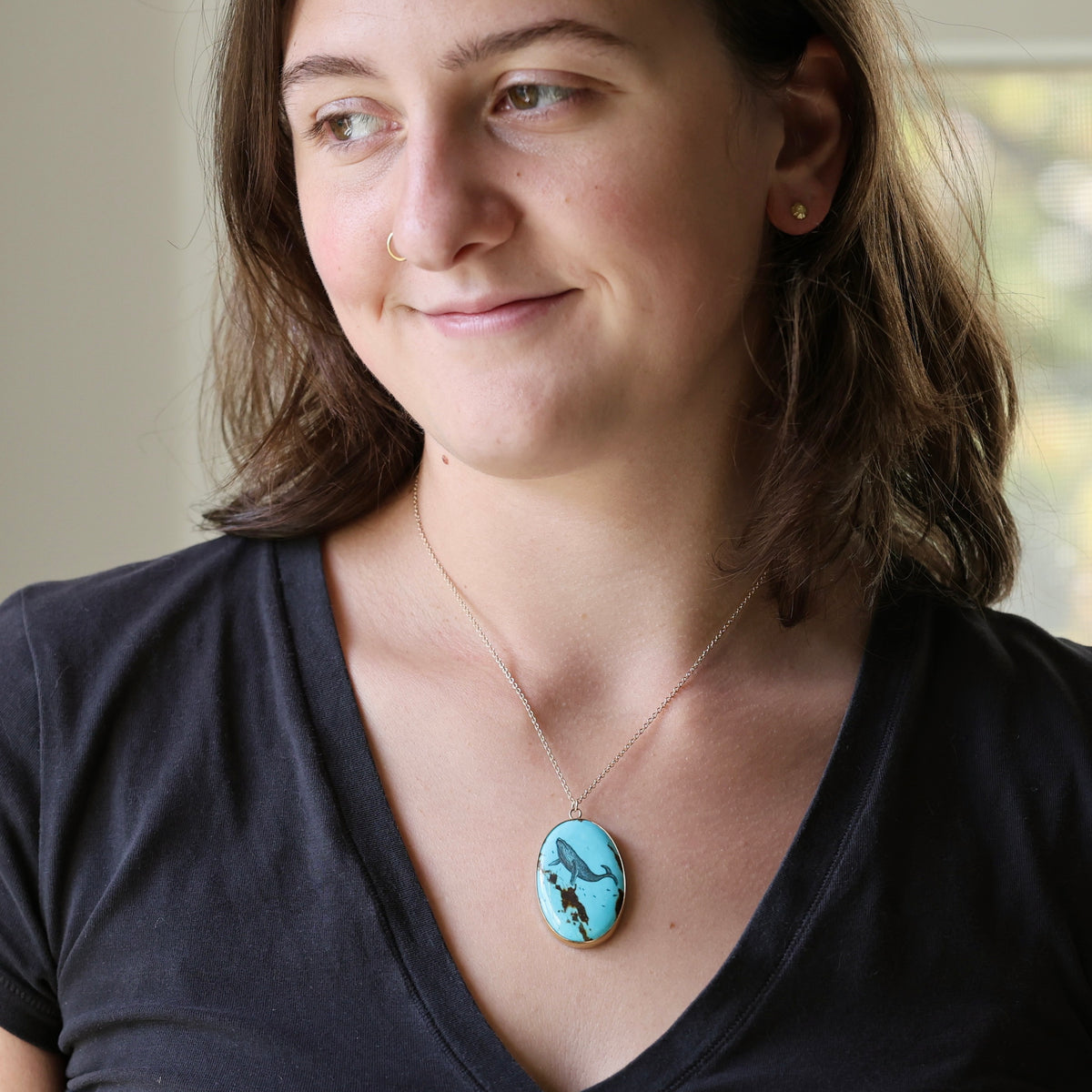 A woman wearing a silver chain necklace with an oval turquoise pendant featuring a black and white graphic design, paired with a black v-neck t-shirt.