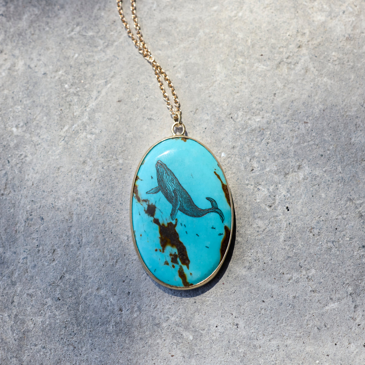 An oval pendant necklace featuring a turquoise stone with a dark blue whale motif and brown matrix, suspended from a gold chain on a textured gray background.