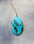 An oval pendant necklace featuring a turquoise stone with a dark blue whale motif and brown matrix, suspended from a gold chain on a textured gray background.