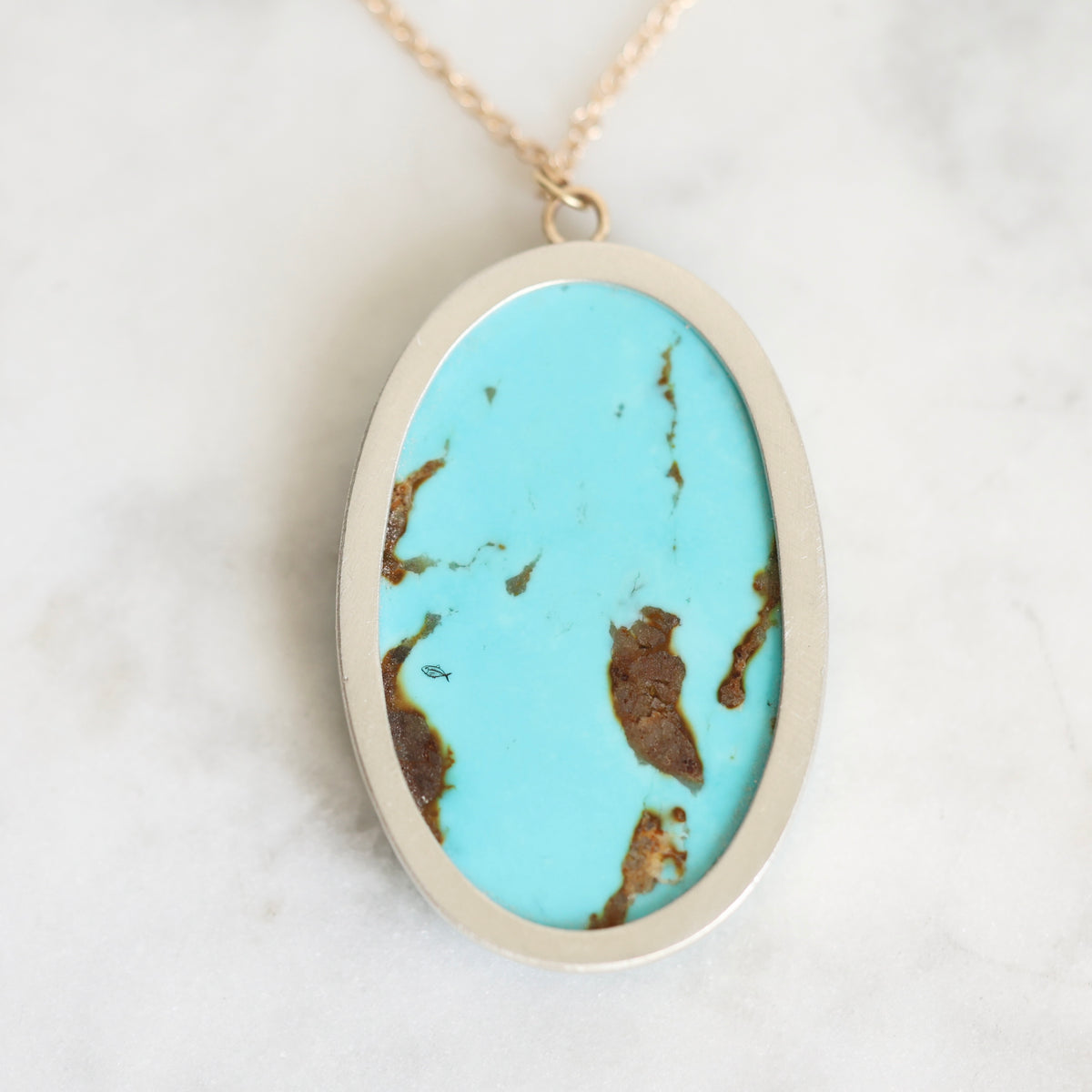 An oval pendant necklace featuring a vibrant turquoise blue stone with natural brown matrix veining, set in a brushed silver-tone metal bezel with a gold-tone chain, resting on a white marble surface.