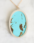 An oval pendant necklace featuring a vibrant turquoise blue stone with natural brown matrix veining, set in a brushed silver-tone metal bezel with a gold-tone chain, resting on a white marble surface.