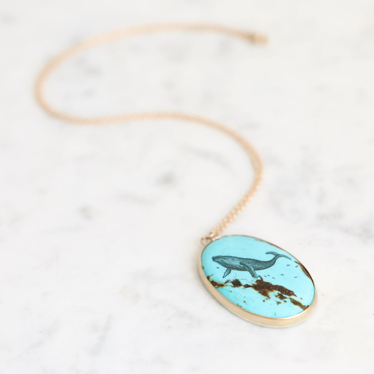 An oval pendant necklace with a light blue turquoise-like background featuring a dark blue whale illustration and brown textured details, suspended from a delicate gold chain on a white marble surface.