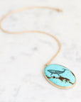 An oval pendant necklace with a light blue turquoise-like background featuring a dark blue whale illustration and brown textured details, suspended from a delicate gold chain on a white marble surface.