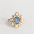 A vintage-style rose gold ring featuring an oval blue gemstone surrounded by a cluster of small rose gold rose-shaped accents and tiny clear stones, set against a plain light gray background.