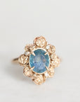 A vintage-style rose gold ring featuring an oval blue gemstone surrounded by a cluster of small rose gold rose-shaped accents and tiny clear stones, set against a plain light gray background.