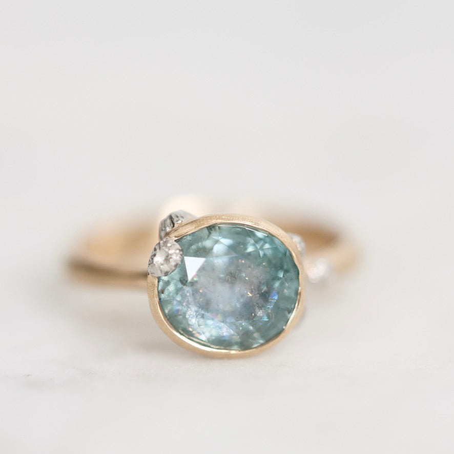 A gold ring featuring a large oval light blue gemstone with tiny inclusions, accented by small clear stones on either side, set against a soft white background.