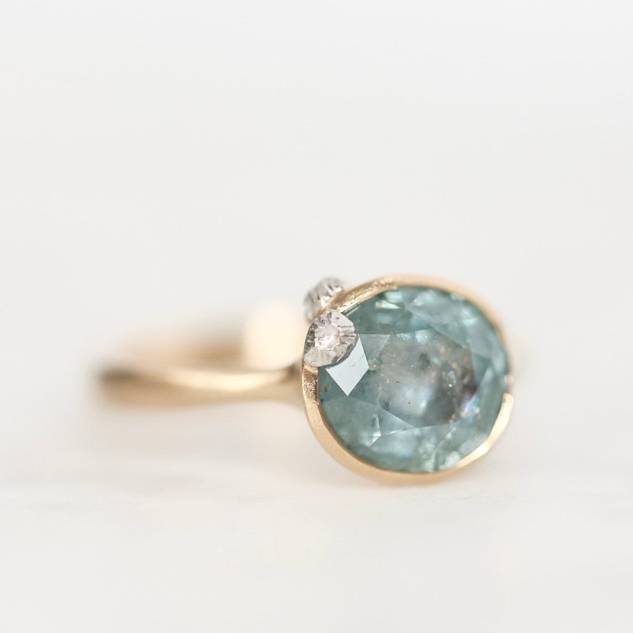 A gold ring featuring a large, round, light blue-green gemstone with a small clear accent stone on the side, set against a clean white background.