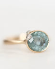 A gold ring featuring a large, round, light blue-green gemstone with a small clear accent stone on the side, set against a clean white background.