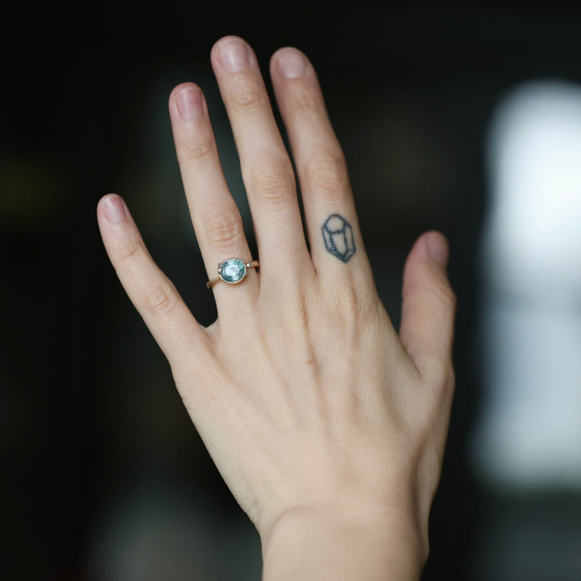 A close-up of a person's hand wearing a ring with a round light blue gemstone in a rose gold setting. The hand also has a small geometric tattoo on one of the middle fingers, and the background is a dark, softly blurred environment.