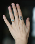 A close-up of a person's hand wearing a ring with a round light blue gemstone in a rose gold setting. The hand also has a small geometric tattoo on one of the middle fingers, and the background is a dark, softly blurred environment.