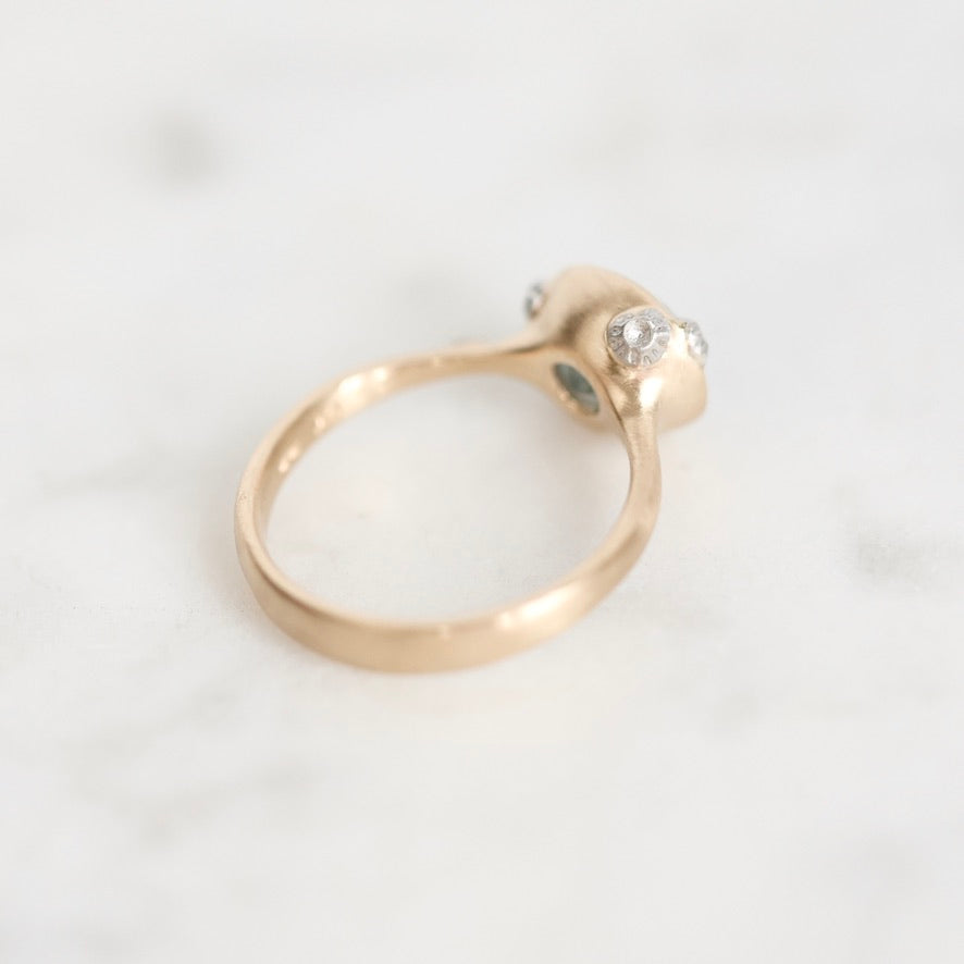 A close-up shot of a gold ring with a unique sculpted setting featuring clear gemstones, resting on a light marble surface.