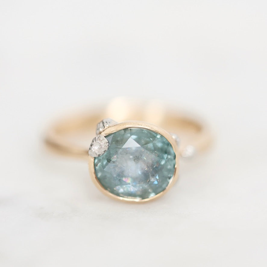 A gold ring featuring a large oval light blue-gray gemstone with small clear accent stones on the shoulders, set against a soft white background.