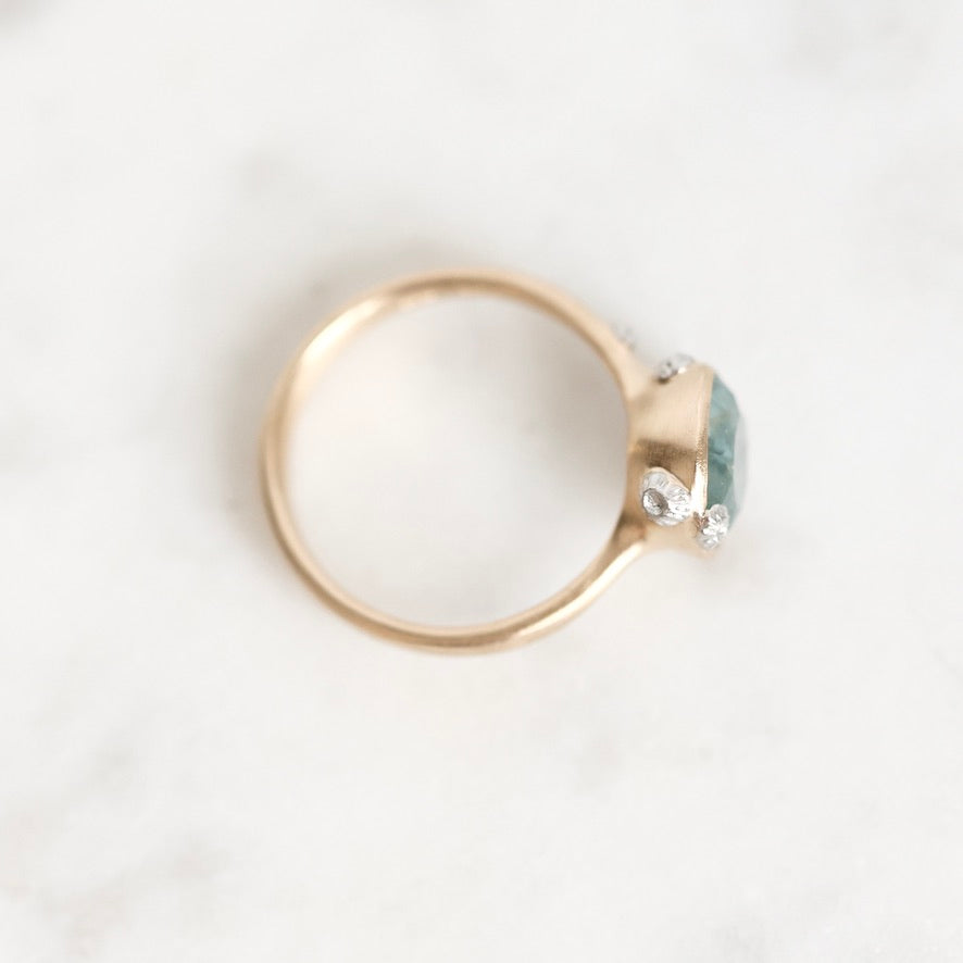 A gold ring with a small oval light blue gemstone and tiny clear accent stones, photographed on a light marble-like surface.