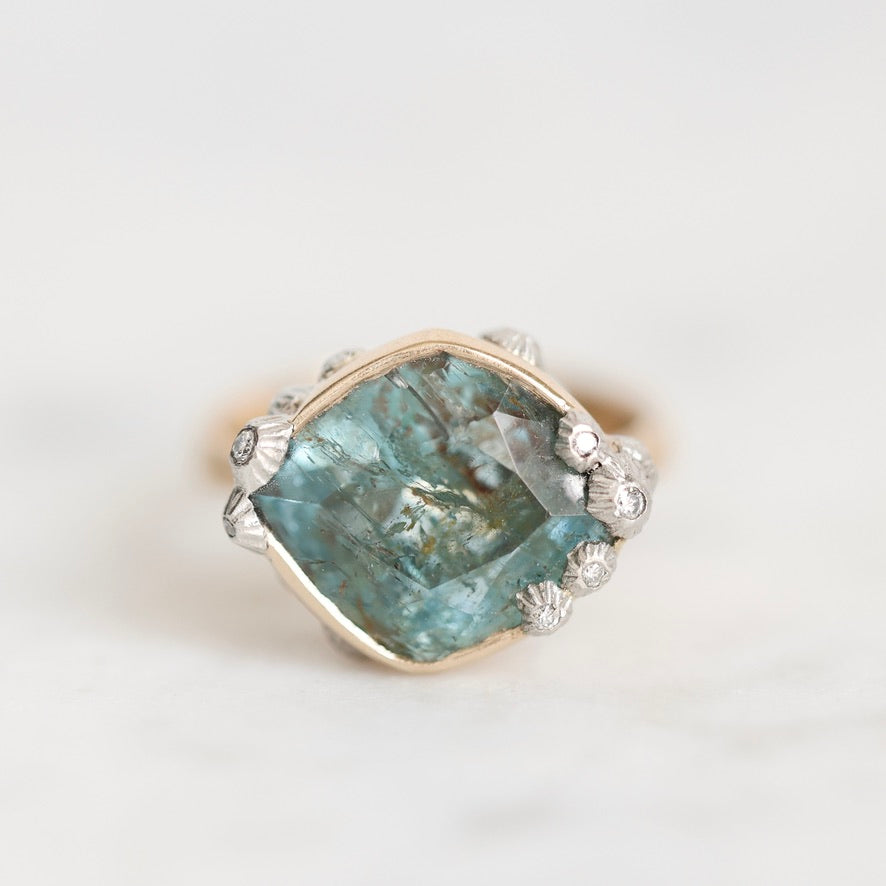 A vintage-style ring featuring a large, rough-cut teal-blue gemstone in a gold-toned setting, accented with small silver-toned floral details and tiny clear stones, set against a clean white background.