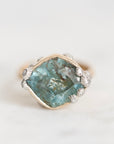 A vintage-style ring featuring a large, rough-cut teal-blue gemstone in a gold-toned setting, accented with small silver-toned floral details and tiny clear stones, set against a clean white background.