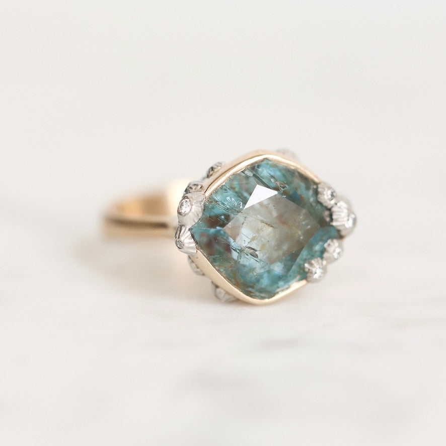 A cushion-cut labradorite ring with a blue-green iridescent center stone, surrounded by a silver halo with small clear accent stones, set on a gold band, photographed on a light marble surface.