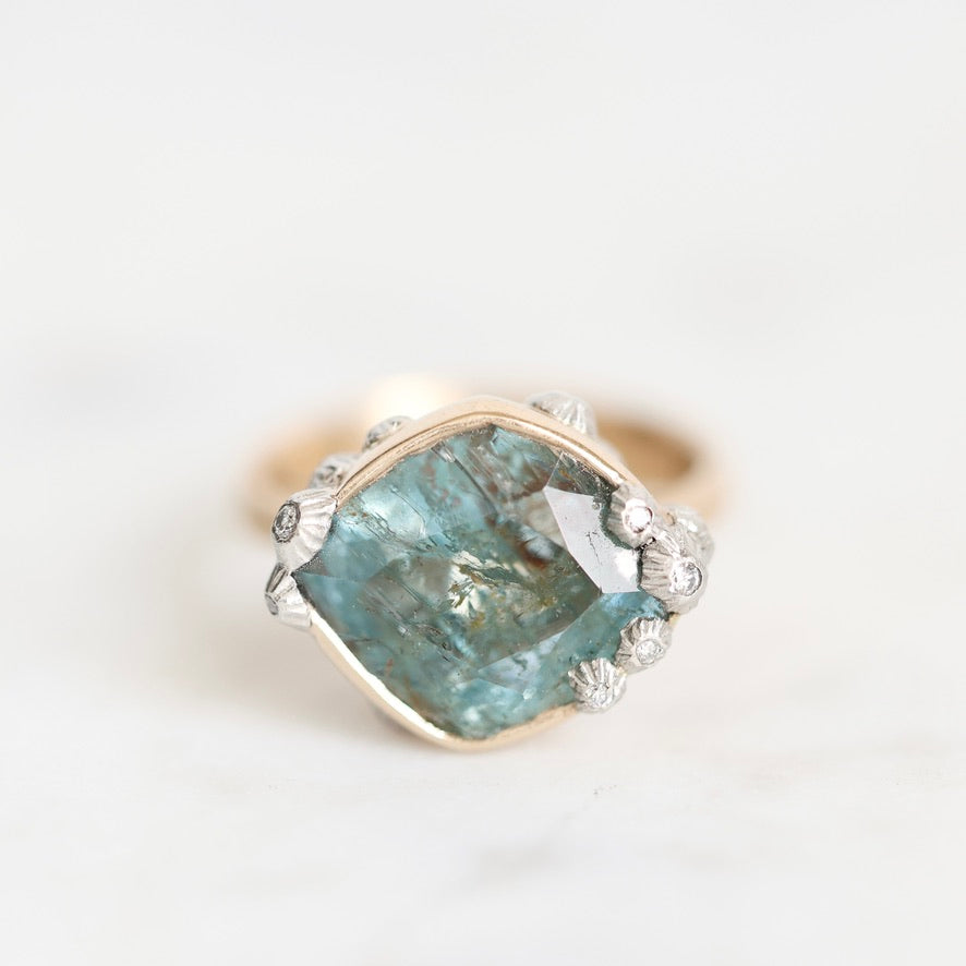 A vintage-style ring featuring a large, irregular-cut light blue gemstone in a gold-toned bezel setting, accented with small silver-toned floral details and tiny clear stones, set against a soft white marble background.