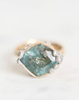 A vintage-style ring featuring a large, irregular-cut light blue gemstone in a gold-toned bezel setting, accented with small silver-toned floral details and tiny clear stones, set against a soft white marble background.