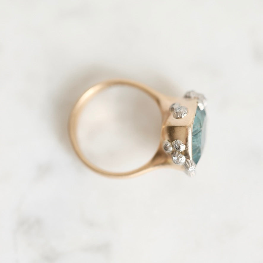 A rose gold ring with a large oval light blue gemstone and small clear accent stones, photographed on a light marble surface.