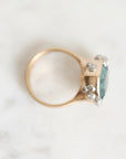 A rose gold ring with a large oval light blue gemstone and small clear accent stones, photographed on a light marble surface.
