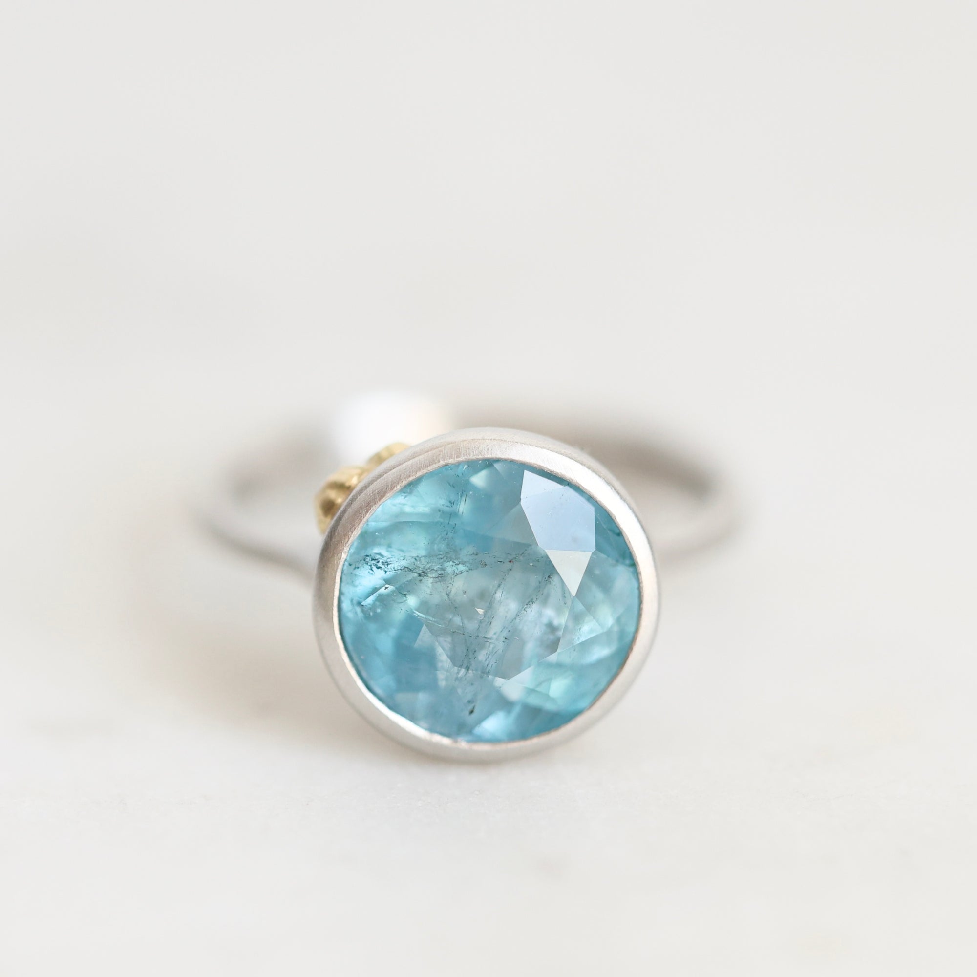 A silver ring featuring a round bezel-set light blue faceted gemstone, photographed on a clean, light off-white background.