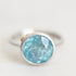 A silver ring featuring a round bezel-set light blue faceted gemstone, photographed on a clean, light off-white background.