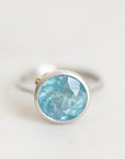A silver ring featuring a round bezel-set light blue faceted gemstone, photographed on a clean, light off-white background.