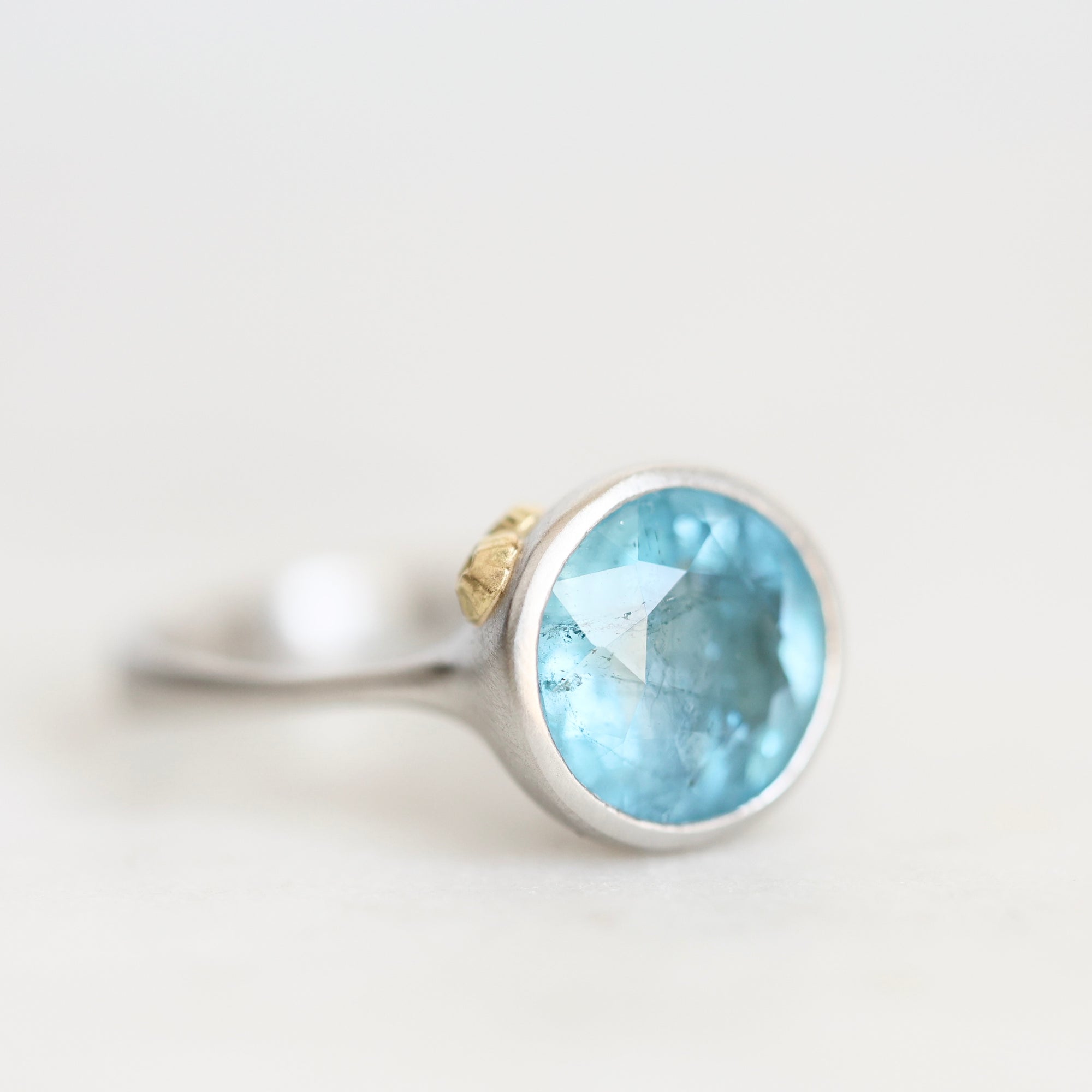 A silver ring featuring a round faceted light blue gemstone, with a small gold accent on the band, set against a plain light background.