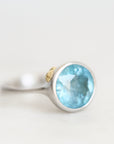 A silver ring featuring a round faceted light blue gemstone, with a small gold accent on the band, set against a plain light background.