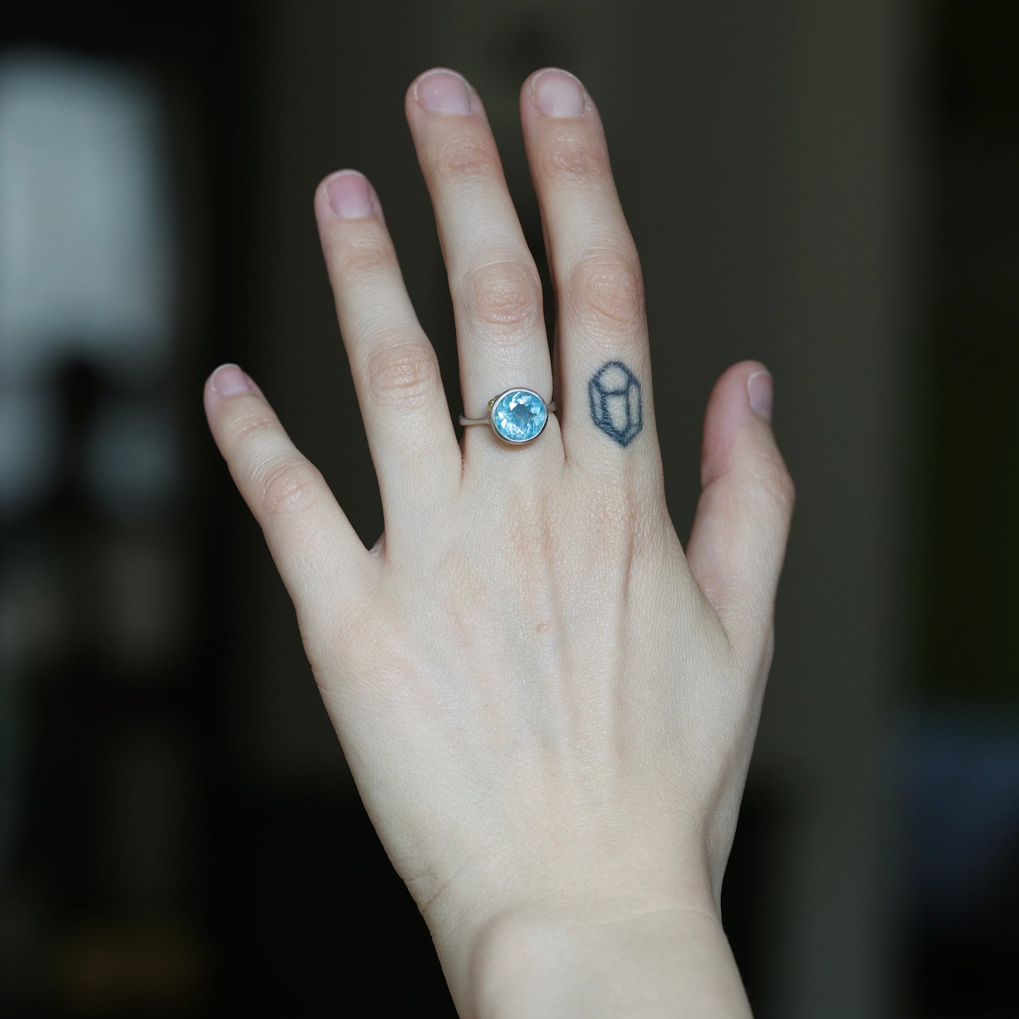 A close-up of a person's hand showing a silver ring with a round light blue gemstone on the ring finger, alongside a small geometric tattoo on the middle finger, against a dark blurred background.