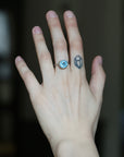 A close-up of a person's hand showing a silver ring with a round light blue gemstone on the ring finger, alongside a small geometric tattoo on the middle finger, against a dark blurred background.