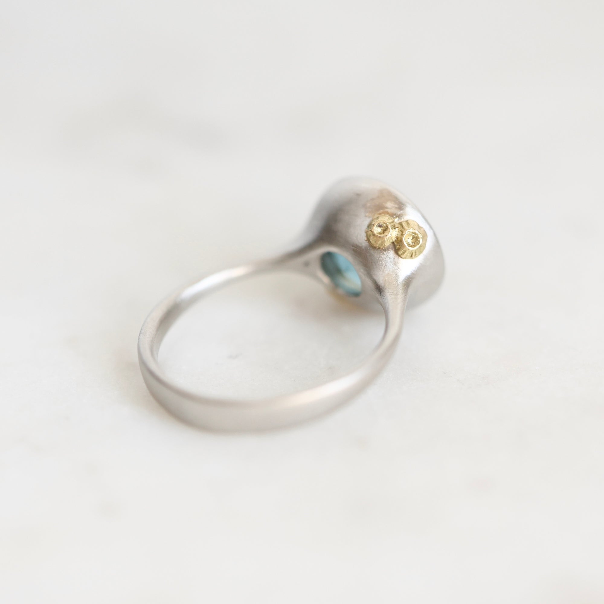 A silver ring with a textured, organic-shaped top featuring two small gold flower-like details and a tiny light blue stone, shown on a light marble surface.
